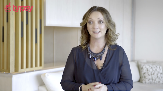 A young woman in a blue dress and necklace presents a Typsy training course in a white room.