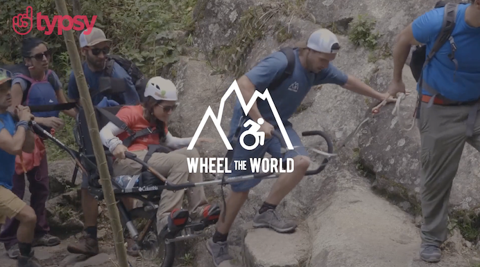A group of people assist a young woman in a wheelchair up a steep, rocky cliff path.